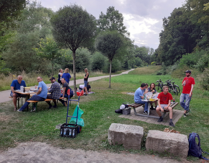 Szene am Grillplatz auf der Wiese am Bach. Menschen sitzen an 
      zwei Tischen, essen und unterhalten sich.